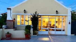 Un edificio con fachada color beige, ventanas grandes y una chimenea de ladrillo. Hay plantas en macetas al frente y un área de entrada pavimentada.