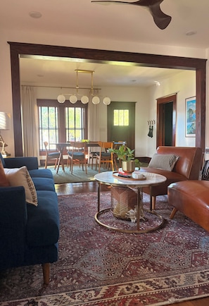 A living room with a blue sofa, a brown leather armchair, a round coffee table, and a dining area with a wooden table and chairs. - Boutique Historic Home • Walk Downtown • Sleeps 6 (New Braunfels)