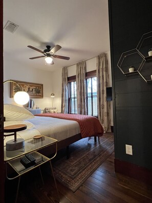 A bedroom with a bed, bedside table, lamp, ceiling fan, window with curtains, and a dark wall with a geometric shelf. - Boutique Historic Home • Walk Downtown • Sleeps 6 (New Braunfels)
