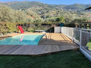 A swimming pool with a wooden deck and a red slide, surrounded by a white fence and a view of a hilly landscape. - Beautiful Villa in Versilia with Pool-Beautiful Villa in Versilia with Pool (Strettoia)