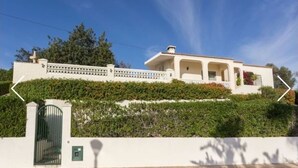 Un bâtiment blanc avec une entrée fermée par un portail, un balcon orné de plantes et un ciel d’un bleu limpide.