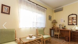 Un salon comprenant un canapé, deux fauteuils, une petite table avec une tasse, une fenêtre avec des rideaux, un bureau, un miroir et un tapis à motifs.