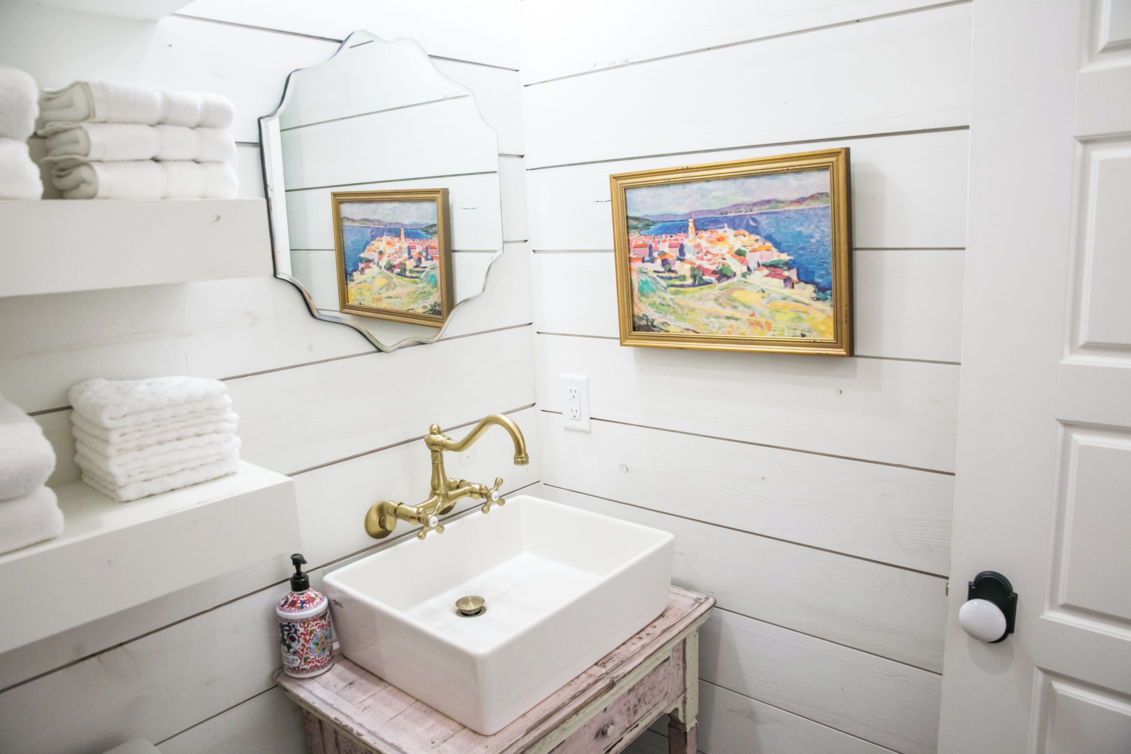 Une salle de bain avec un lavabo blanc, un robinet doré, un miroir et deux tableaux encadrés accrochés au mur.