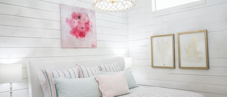 Ein Schlafzimmer mit weißer Holzvertäfelung, einem Bett mit gestreifter Bettwäsche, einem rosafarbenen abstrakten Gemälde und zwei gerahmten Pflanzenbildern.
