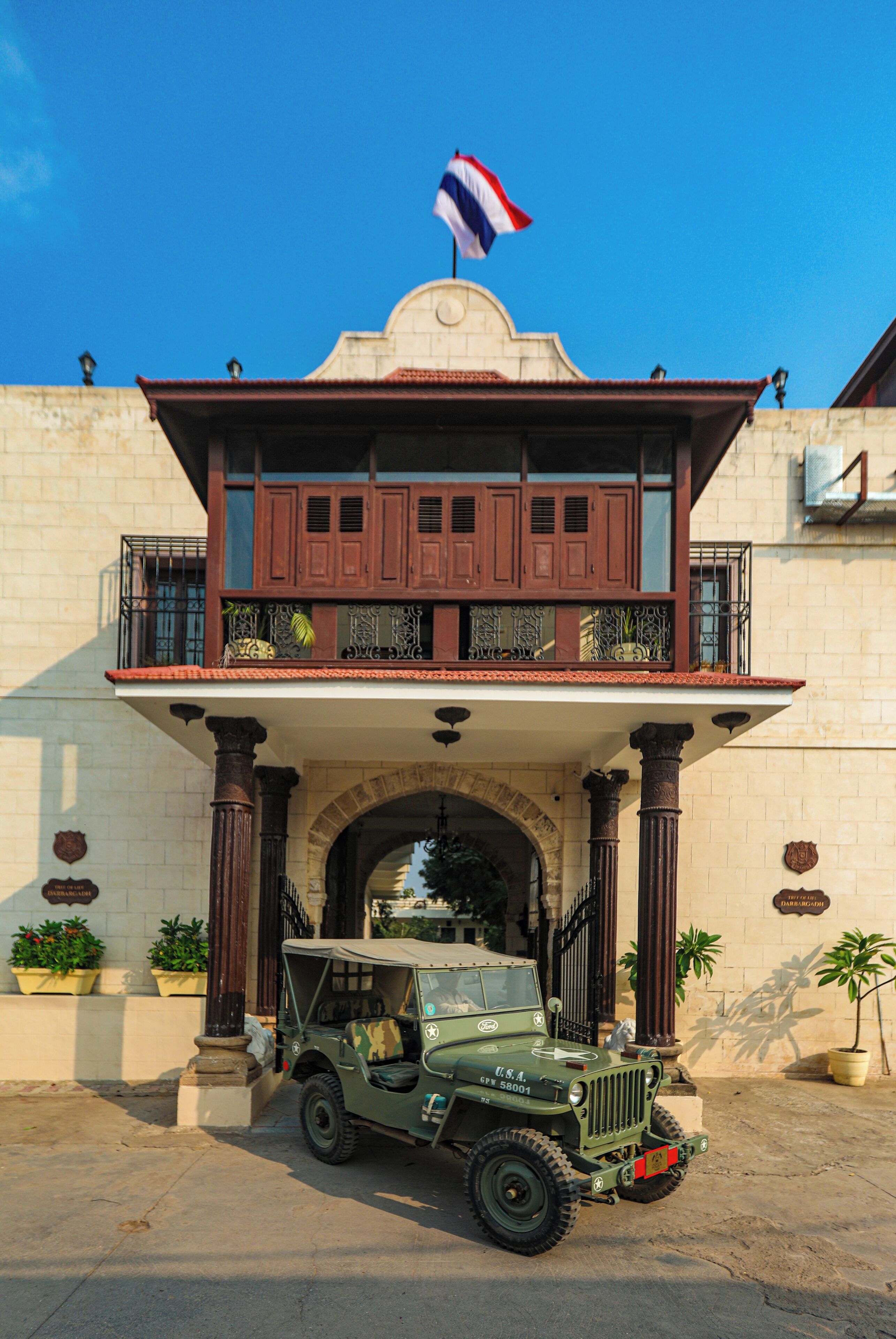 Un edificio de estilo colonial con un balcón, un jeep estacionado adelante y una bandera en la cima.