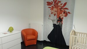 A room with a white dresser, an orange chair, a crib, and a large floral artwork on the wall.