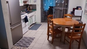 Una cocina con refrigerador, estufa y alacenas. Hay una mesa de comedor con sillas y un tapete estampado.