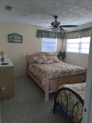 A bedroom with a bed, a desk, a ceiling fan, a window with blinds, and a wall-mounted sign.