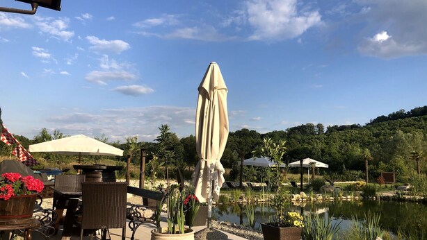 Uma área externa com mesas, cadeiras, um grande guarda-sol e um lago rodeado por vegetação.