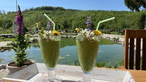 Duas taças de bebida verde com canudos e flores sobre uma bandeja de madeira, junto a um lago.