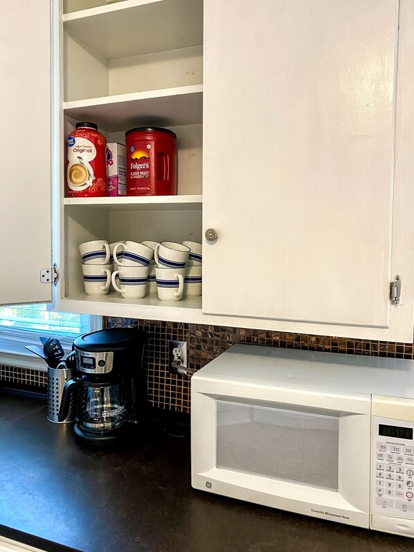 A kitchen with a coffee maker, a microwave, and a cabinet containing coffee cups and Folgers coffee.