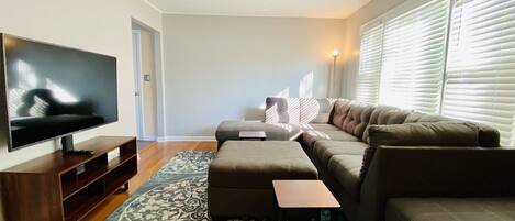 A modern living room with a flat-screen TV, a sectional sofa, a small ottoman, and a patterned rug.