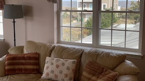 A living room with a beige sofa, a floor lamp, and a large window offering a view of a residential area and the ocean. - Steps From The Beach and Minutes to The Boards - Now Includes Linens (Milton)