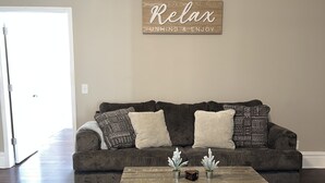 Uma sala de estar com piso de madeira marrom escuro, um sofá cinza com almofadas estampadas e uma mesa de centro rústica com objetos decorativos. Um aviso na parede diz "Relax Unwind & Enjoy".