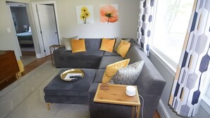 Ein modernes Wohnzimmer mit einem Ecksofa, einem kleinen Holztisch und Blick auf einen Garten durch bunte Vorhänge.