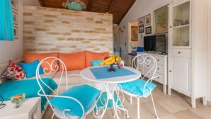 Un cuarto acogedor con pared de piedra, un sofá blanco con cojines anaranjados, una mesa redonda con frutero y un armario blanco con puertas de cristal.