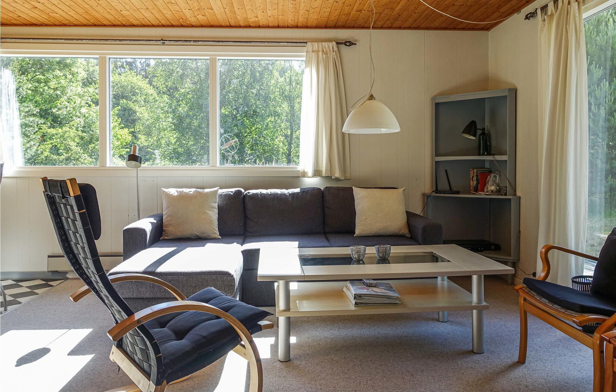 Una sala de estar acogedora con un sofá, una mesa de centro y un sillón mecedora. Se ve una ventana con cortinas y una estantería al fondo.