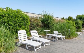 Des fauteuils de jardin et une petite table sont disposés sur une surface pavée, avec en arrière-plan une végétation luxuriante et un bâtiment visible au loin.