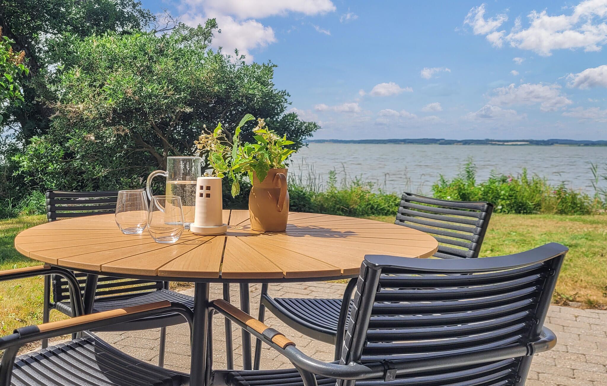 An outdoor table with chairs, a pitcher, and glasses, set against a backdrop of a lake and trees.