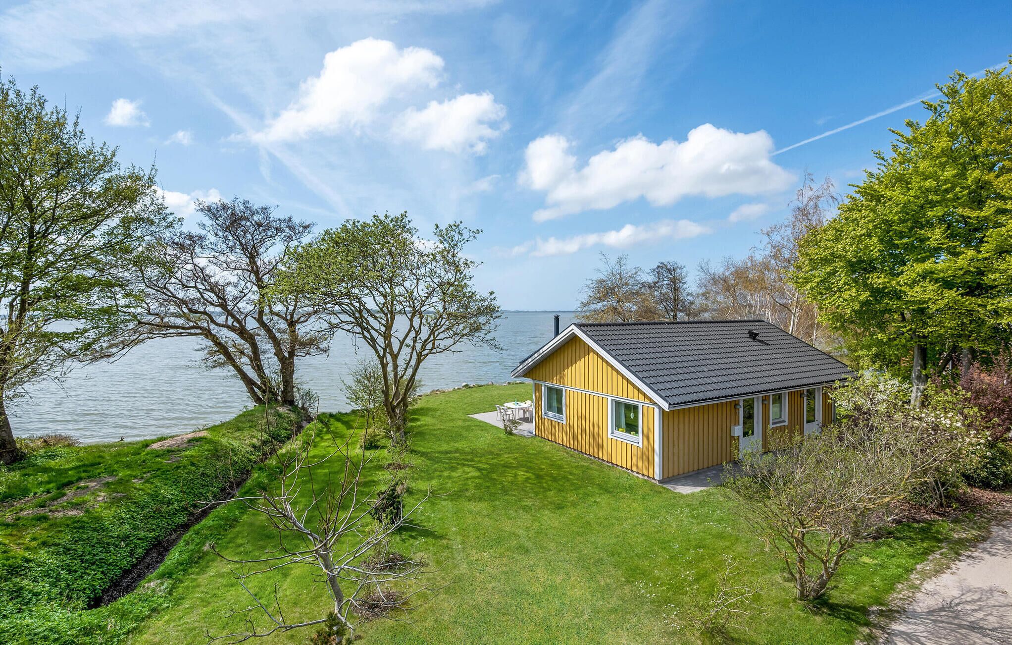 A yellow cabin with a black roof, surrounded by a grassy area and trees, near a body of water.