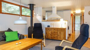 A cozy living room with a wooden table, a sofa with a green pillow, and a black chair. A kitchen area is visible in the background with wooden cabinets and a white stove. - Amazing home in Nexø with WiFi (Nexø)