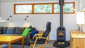 A cozy living room with a sofa, a wooden table, a rocking chair, a wood-burning stove, and a window with a view of trees. - Amazing home in Nexø with WiFi (Nexø)