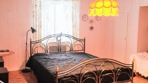 Une chambre à coucher avec un lit en métal, une table de chevet, une lampe et une fenêtre ornée de rideaux à motifs floraux.
