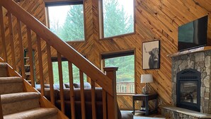 A wooden interior with a staircase, a stone fireplace, and large windows.