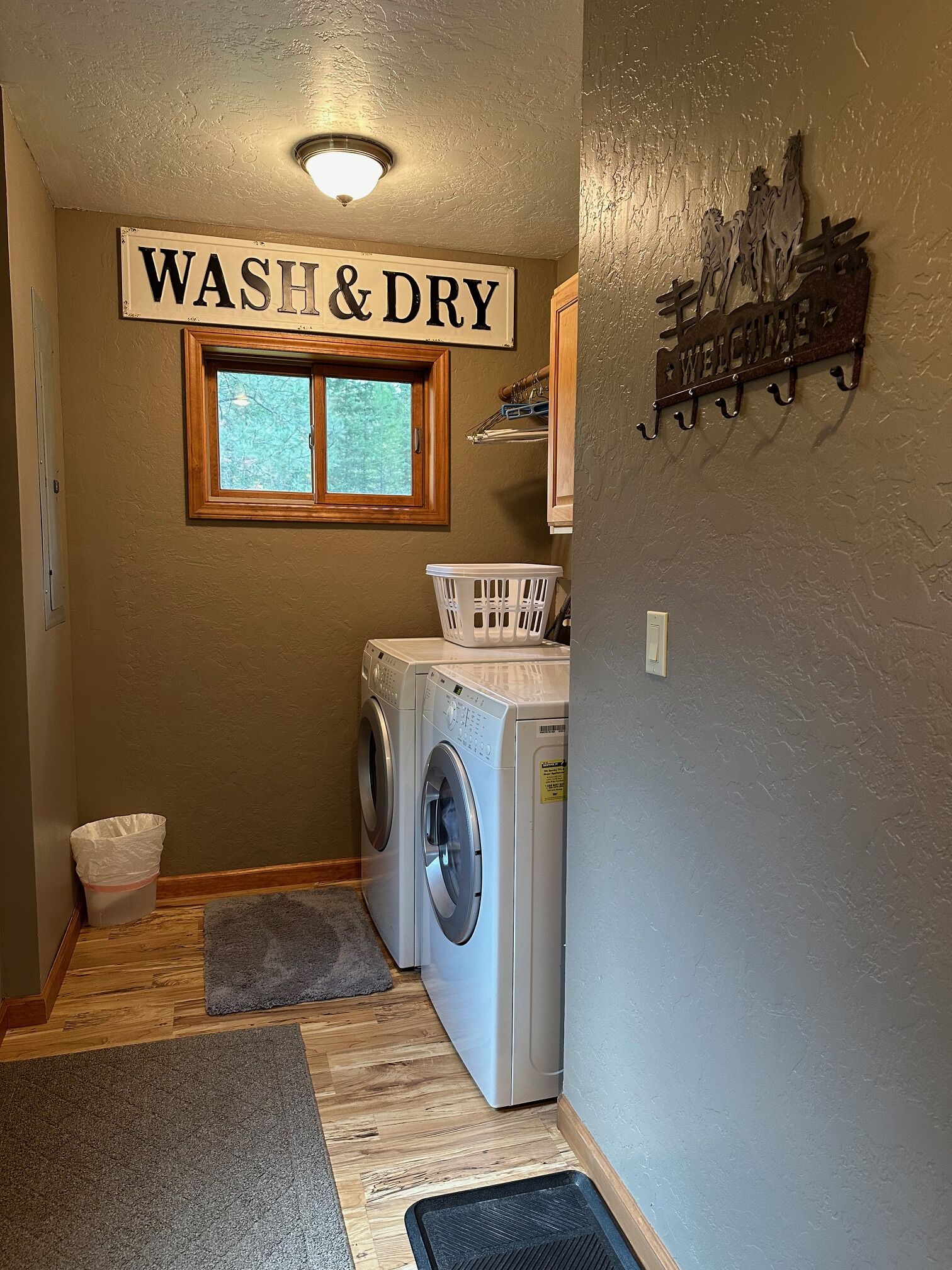 A laundry room with a 'WASH & DRY' sign, a washing machine, a dryer, a window, a welcome sign, and a small rug on the floor.