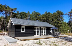 Eine kleine, dunkle Holzhütte mit einem Ziegeldach, einer Terrasse mit Gartenmöbeln und einem sandigen Strand im Hintergrund.