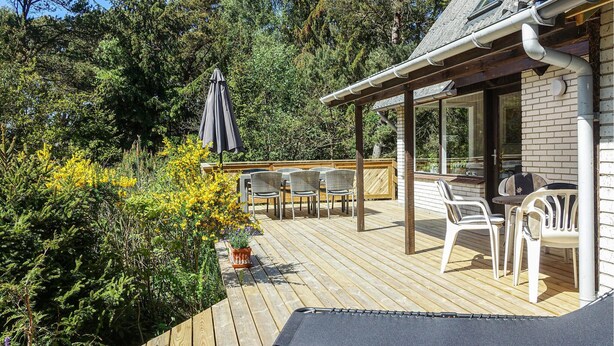 Eine Holzterrasse mit Gartenmöbeln, einem überdachten Sitzbereich und Blick auf einen bewaldeten Bereich.