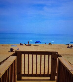 Ein Holzsteg führt zu einem Strand mit Menschen und Sonnenschirmen.