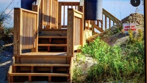 Un escalier en bois mène à une zone de plage où se trouvent deux structures cylindriques, qui pourraient être des poubelles, ainsi qu’un panneau indiquant : « IL EST INTERDIT DE MARCHER SUR LES DUNES ».