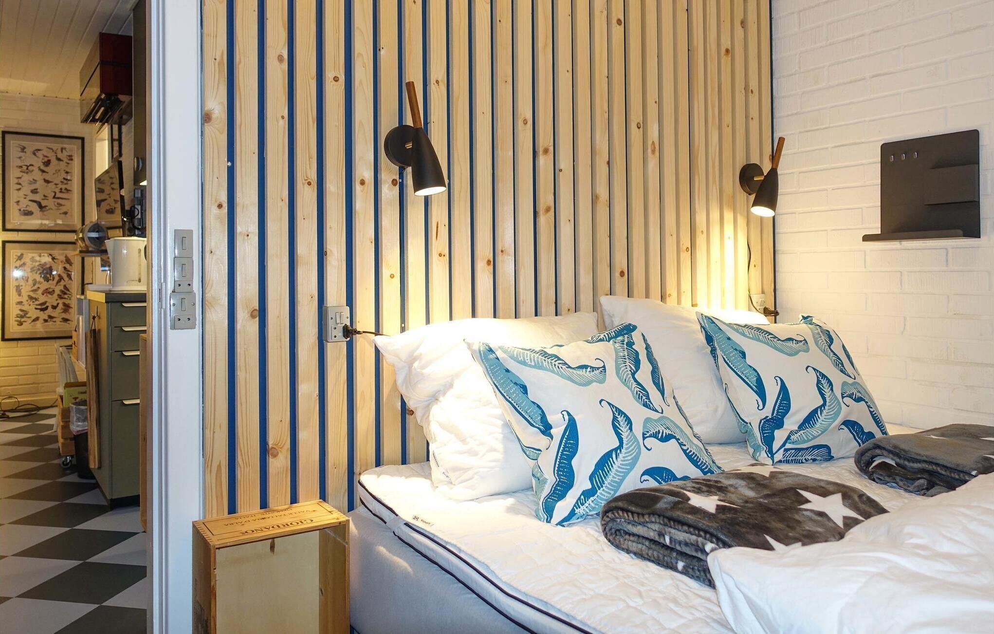 A bedroom with a wooden slat wall, a bed with white bedding and blue leaf-patterned pillows, and two wall-mounted reading lamps.