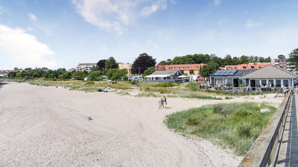Uma praia com areia, algumas pessoas e edifícios ao fundo.