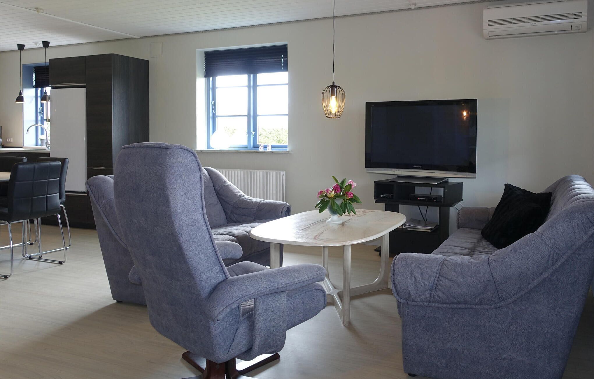 Ein modernes Wohnzimmer mit Flachbildfernseher, grau gepolsterten Sesseln, einem kleinen weißen Tisch und einer Pendelleuchte.