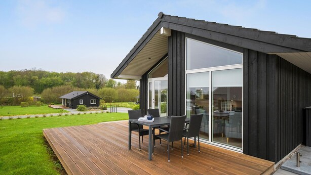 A modern house with a wooden deck, a dining area with chairs, and a glass door leading to a garden. - 2 bedroom gorgeous home in Haarby (Haarby)