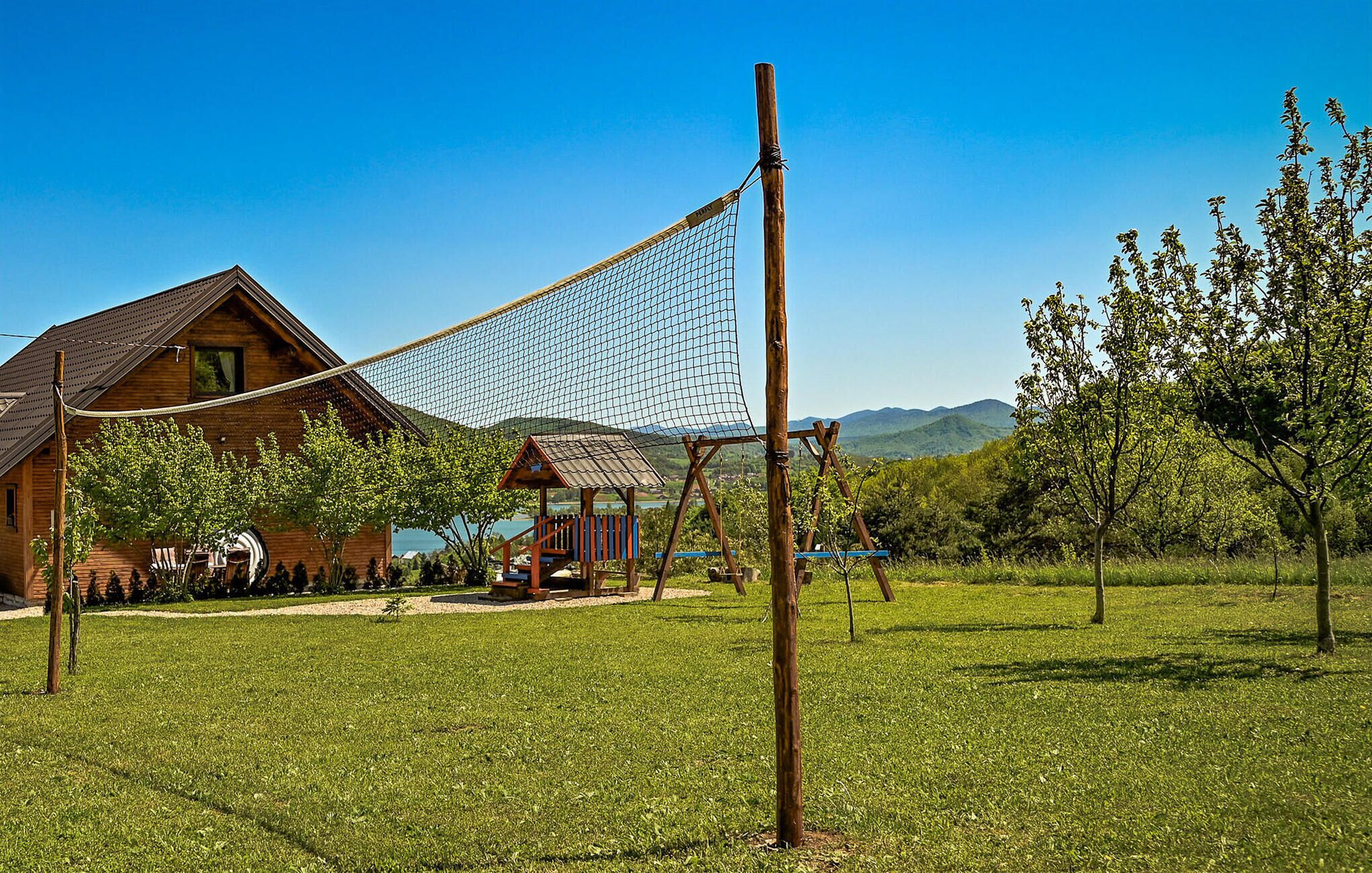 Une maison en bois avec un court de tennis, située dans un champ verdoyant et entourée d’arbres et de montagnes.