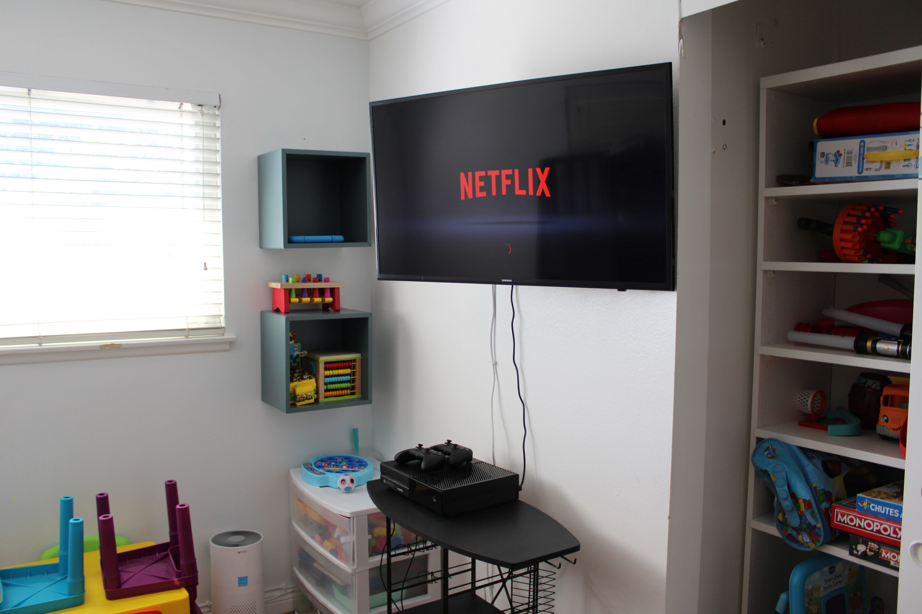 Una habitación con un televisor en la pared mostrando el logotipo de Netflix, una mesita con un altavoz y un mando a distancia, y estantes con juguetes y juegos.