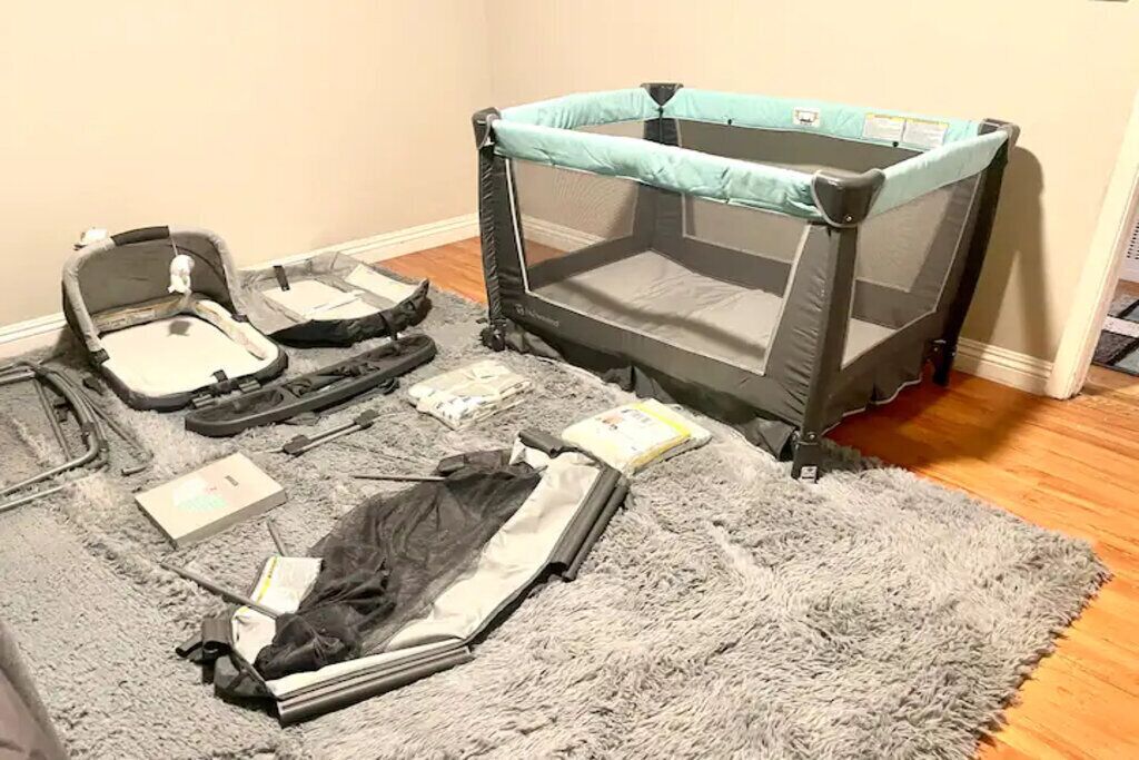 A room with a grey furry rug, a grey and black baby playpen, and scattered baby items.