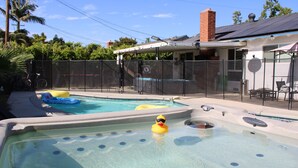 A backyard with two swimming pools, one with a hot tub, surrounded by a fence, and a house in the background.