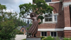 En statue af en kvinde med udstrakte arme på en rund fod, mærket "POLLYAN", foran en murstensbygning.