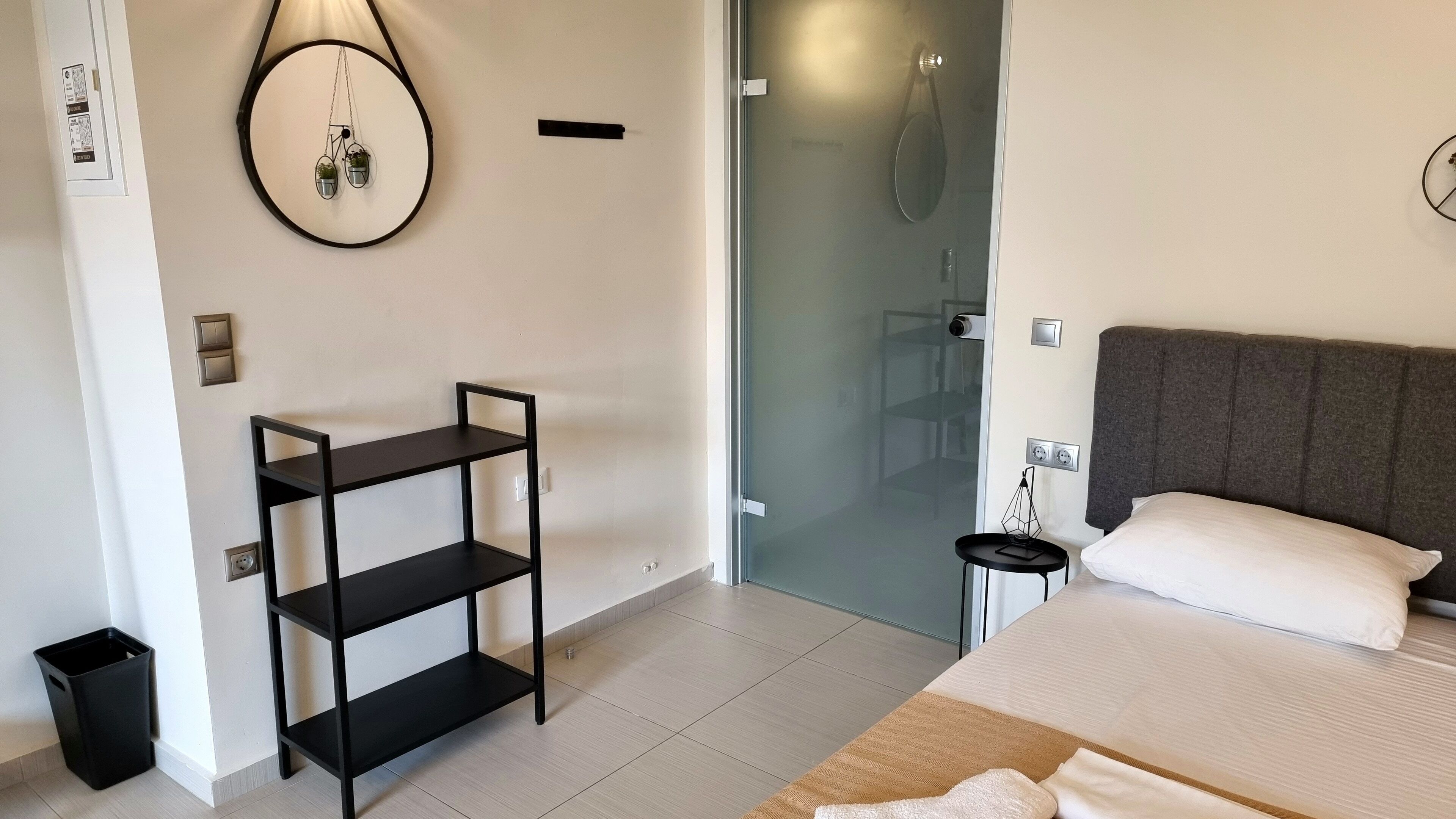 Una camera d'albergo con un letto, un piccolo ripiano nero, una cabina doccia in vetro e un lampadario a parete.
