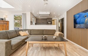 A modern living room with a sectional sofa, a coffee table, and a flat-screen TV mounted on a wooden wall. - 4 bedroom nice home in Nexø (Nexø)
