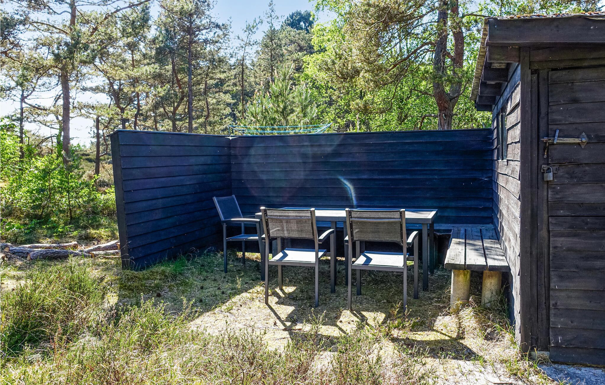 Et overdækket udendørs opholdsområde med et bord og stole, omgivet af et træhegn og beplantning.