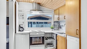 Cocina compacta con estufa, horno y cafetera. Incluye un ventanal con vista a una cerca azul y un techo de madera.
