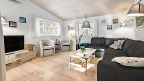 Ein modernes Wohnzimmer mit Flachbildfernseher, einer Couch, einem Couchtisch und einem Esstisch. Zwei Stühle, eine Lampe und ein Fenster mit Vorhängen sind ebenfalls vorhanden.
