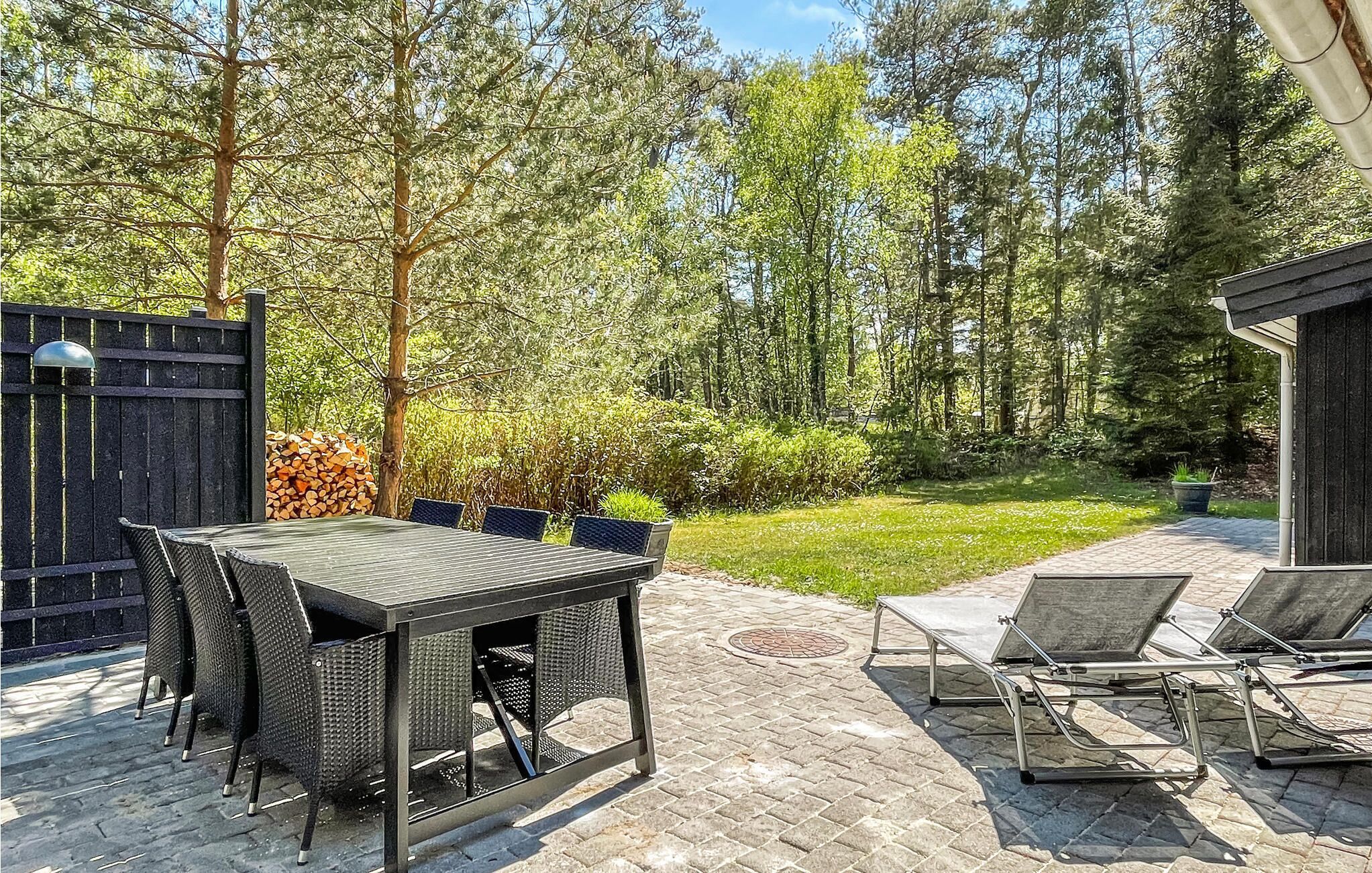 An outdoor patio with a table and chairs, surrounded by a wooden fence and a stack of firewood.