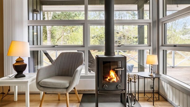 Una habitación acogedora con un amplio ventanal, un sillón cómodo, una mesa con lámpara y un estufa de leña.