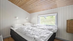 Un dormitorio con techo de madera, paredes blancas, un ventanal grande y una cama con ropa blanca.